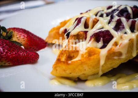 Nahaufnahme von goldenen Waffeln, die mit cremiger weißer Sauce und dunklem Beerenkompott überzogen sind, serviert mit frischen Erdbeeren auf einem weißen Teller. Stockfoto