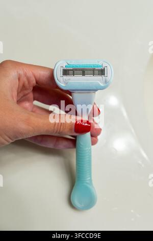 Hand mit rotem Nagellack, die einen blauen Mehrklingen-Plastikrasierer über einer hellen Badezimmeroberfläche hält. Rutschfester ergonomischer Griff, scharfe Klingendetails Stockfoto