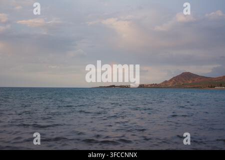 Ruhige Seelandschaft mit sanften Wellen unter einem bewölkten Himmel und einem schwachen Regenbogen über der fernen bergigen Küstenlinie in der Dämmerung. Stockfoto