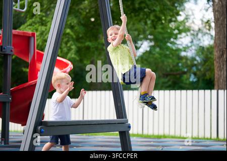 Junge klettert an einer Seilbahn auf dem Spielplatz, während ein anderer Junge unten zusieht, aktives Spielen im Freien, Parkumgebung, Kindheitsabenteuer und Spaß Stockfoto