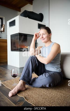 Junge Frau verbringt einen ruhigen Abend am Kamin, entspannt mit einem Getränk in einem modernen, gemütlichen Zuhause, Lifestyle- und Komfortkonzept Stockfoto