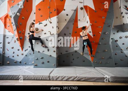 Zwei junge Frauen bouldern an einer farbenfrohen Indoor-Kletterwand, beide in schwarzer Sportkleidung. Seiten- und Rückansicht, Sturzmatten unten. Ideal für Fitness-, Teamwork- und Active-Lifestyle-Werbung Stockfoto
