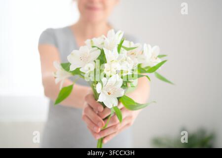 Eine Frau hält einen weißen Blumenstrauß in ihren Händen in einem hellen Raum. Die Dame, die Blüten wiegt, präsentiert einen ruhigen, friedlichen Moment zarter nachdenklicher Gelassenheit und reiner, zarter Schönheit im Innenraum. Stockfoto
