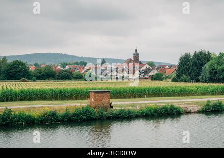 Ländliche Aussicht über Ackerland in Richtung des Dorfes Kleinheubach in Bayern, Deutschland, mit einem markanten Kirchturm unter einem bewölkten Sommerhimmel. Stockfoto