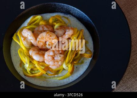 Oberansicht von gelben spiralisierten Zucchininudeln mit gebratenen Garnelen auf cremiger Kräutersauce in einer dunklen matten Schale auf gewebtem braunem Textil Stockfoto