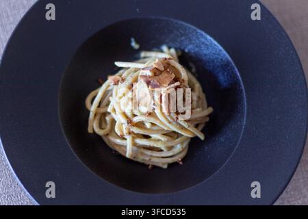 Oberansicht eines Vollkornspaghettinests mit cremiger Sauce, gerösteten Mandelblättchen und Paniermehl in einer schwarzen Schüssel auf beigem Leinen Stockfoto