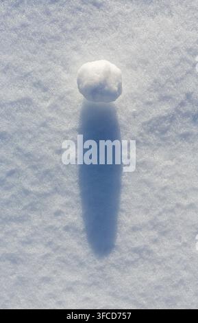 Ein einzelner weißer Schneeball wirft einen langen blauen Schatten auf strukturiertem Schnee im Abendlicht. Stockfoto