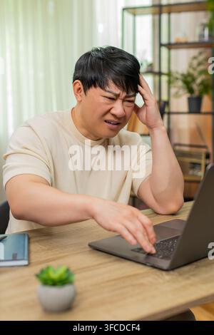 Asiatischer Mann, der am Tisch sitzt, liest schlechte E-Mails auf dem Laptop erneut, klopft wütend auf die Tastatur, die Stirn tief gefurcht. Der Chinese zeigt eine angespannte Frustration, die Schultern sind starr, Enttäuschung und Stress sichtbar. Stockfoto