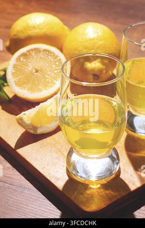 Zwei klare Gläser stehen auf einem Holzbrett, das mit gelbem Zitronentrink gefüllt ist. Ganze und geschnittene Zitronen sind um die Gläser auf dem Tisch angeordnet Stockfoto