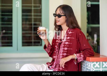 Stylische indonesische Frau trägt eine moderne Batikbluse und eine Sonnenbrille und genießt einen Kaffee, während sie in einem farbenfrohen Café-Interieur entspannt. Stockfoto