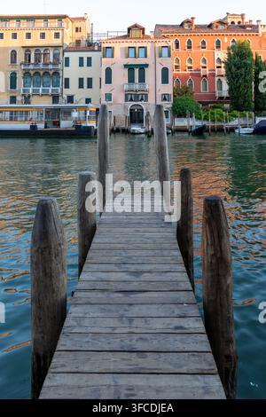 Holzsteg an einem venezianischen Kanal mit farbenfrohen historischen Häusern am Ufer, ruhigem, welligem Wasser und einem kleinen Boot an einem italienischen Stadtabend. Stockfoto