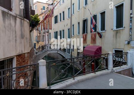 Ruhiger schmaler Kanal in Venedig, Italien, mit einer gewölbten Stein-Fußgängerbrücke, alten pastellfarbenen Gebäuden, eisernen Geländern und kleinen Booten neben dem Wasser. Stockfoto