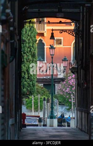 Blick durch eine dunkle, enge Gasse in Venedig auf einen Kanal, alte Straßenlaternen, Blumen und ein warmes, orangefarbenes historisches Gebäude in Italien. Stockfoto