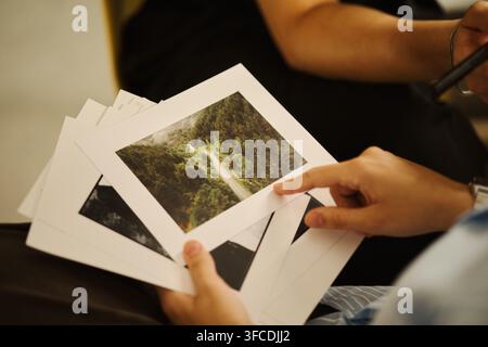 Hände eines jungen asiatischen Mannes, der gedruckte Landschaftsfotos während einer kreativen Portfoliobesprechung hält und darauf zeigt, repräsentierend Fotografiegeschäft und künstlerische Kuratierung, Nahaufnahme Stockfoto