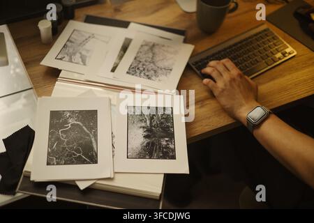 Männlicher Fotograf tippt mit der Hand auf der Tastatur neben gedruckten Schwarzweiß- und Kunstfotos, organisiert Portfolio. Bild geeignet für kreative Agentur oder Kunstgaleriemarketing Stockfoto