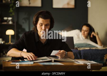 Junger asiatischer Mann betrachtet gedruckte Fotos in einem Album, während sich eine junge Frau im Hintergrund auf dem Bett entspannt. Das Bild steht für Nostalgie und die Bewahrung von Erinnerungen für die Familienhistorie-Marketing Stockfoto