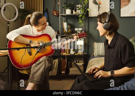 Kaukasische junge Frau spielt Akustikgitarre, während kaukasischer junger Mann mit Kopfhörern Laptop verwendet. Kreatives Duo nimmt Musikpodcast auf, geeignet für Home-Studio-Marketing Stockfoto