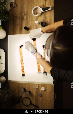 Hohe Blickwinkelaufnahme einer jungen kaukasischen Frau, die Handschuhe trägt und Analogfilmstreifen auf einem Lichttisch untersucht, was fotografische Archivierungs- und Restaurierungsdienste repräsentiert, vertikal Stockfoto