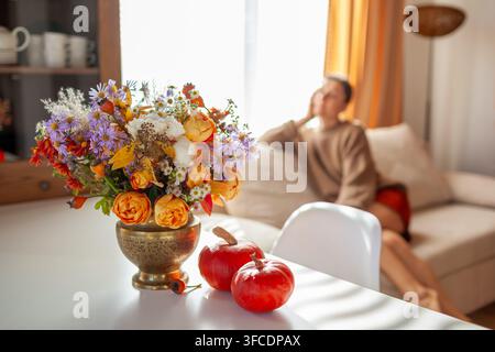 Herbstliche Inneneinrichtung eines Wohnzimmers. Frau entspannt sich zu Hause, das für den Herbst mit einem Blumenstrauß, orangefarbenen Kürbissen dekoriert ist. Gemütliches Erntedankfest Stockfoto
