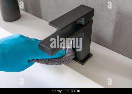 Eine Person, die einen blauen Schutzhandschuh trägt, wischt einen glatten schwarzen Badezimmerwasserhahn mit einem grauen Mikrofasertuch ab und betont Hygiene und Sauberkeit Stockfoto