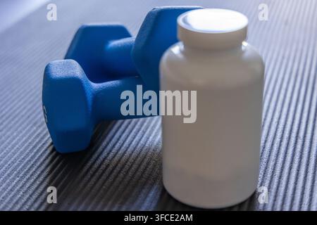 Ein Paar helle blaue Hanteln ruht neben einer weißen undurchsichtigen Flasche mit Nahrungsergänzungsmitteln auf einer dunklen gerippten Matte, was auf eine Fitness-Routine hindeutet Stockfoto