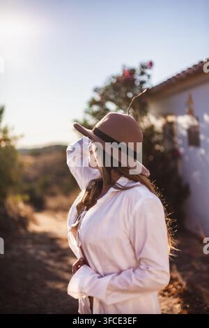 Junge Frau in weißem Kleid, barfuß auf einer ländlichen Straße sitzend, hält einen großen hölzernen Bilderrahmen bei Sonnenuntergang, Algarve, Portugal Stockfoto