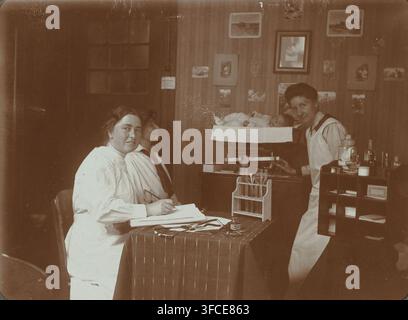 Charlotte van der Schilt Ausbildung zur Kinderärztin, aus dem Album Photographien, Albumseite, Fotografie, Dieses Foto ist Teil eines Albums., Fotograf: zugeschrieben Christiaan Otto Roelofs, Amsterdam, 1908, Zugeschrieben, Papier, Tageslicht-Gelatine-Silberdruck, Höhe 80 mm x Breite 110 mm, Person, Charlotte Roelofs-van der Schilt, historische Personen - BB - Frau, Ärztin, Doktor., Eine Sepiafotografie, die einen Raum mit gemusterter Tapete und einer Sammlung gerahmter Bilder an der Wand zeigt, ein Tisch, der mit einem gestreiften Tuch bedeckt ist und Papiere, Instrumente, eine Tintenflasche und ein hölzernes Gestell hält Stockfoto