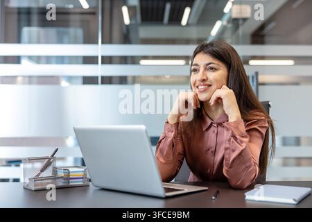 Junge Berufsfrau lächelt und schaut weg, während sie an ihrem Schreibtisch in einem modernen Firmenbüro sitzt und über Geschäftserfolge und zukünftige Ziele nachdenkt Stockfoto