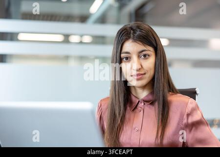 Junger Geschäftsmann posiert in einer modernen Büroumgebung, der Kamera mit einem selbstbewussten Ausdruck gegenübersteht, während er einen Laptop nutzt, und somit unternehmerischen Erfolg und Karriereentwicklung repräsentiert Stockfoto