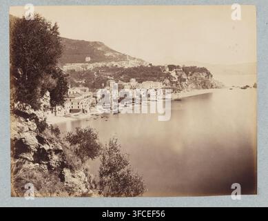 Ansicht der Küste von Salerno, Contorni di Salerno. Vietri strada di Amalfi, Fotografie, Capri, Isola di, 1851 - 1900, Fotograf, Karton, Papier, Albumen-Druck, Höhe 192 mm x Breite 250 mm, Höhe 251 mm x Breite 320 mm, Küste, Stadtansicht im Allgemeinen, 'veduta'. Eine Küstenszene, die ein Hügeldorf mit einer Ansammlung hellfarbener Gebäude zeigt, die sich hinunter zu einem schmalen Strand hinabschlingen, an dem mehrere kleine Boote am Ufer liegen und einige Boote in dem ruhigen Wasser ruhen; steile felsige Klippen und Vegetation rahmen die linke Seite des Bildes mit Bäumen und Sträuchern, die auf dem Hang sichtbar sind, und der Stockfoto