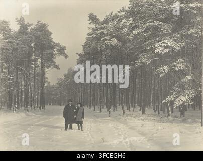 Mann und Mädchen im Schnee auf dem Boslaan im Berg en Bosch Park in Apeldoorn, Schneeiger Boschlaan im Berg en Bosch Park in Apeldoorn, Fotografie, Anmerkung: 'Schneeiger Boschlaan, im Berg en Bosch Park, in Apeldoorn', Signatur: 'Foto, R Tepe', Fotograf: Richard Tepe, Niederlande, 1910 - 1930, Papier, Gelatinsilberdruck, Entwicklung Gelatinsilberdruck, Höhe 168 mm x Breite 223 mm., Eine verschneite Waldszene mit hohen Kiefern, die mit Schnee bedeckt sind und sich in den Hintergrund erstrecken, und ein Mann und eine Frau, die im Vordergrund auf einem schneebedeckten Pfad stehen; der Mann trägt einen langen dunklen Mantel und Hut und Stockfoto
