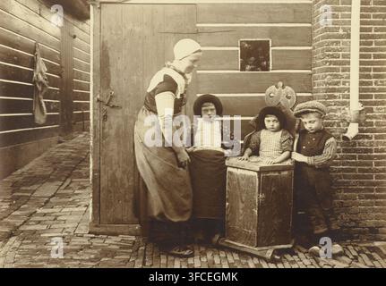 Frau und drei Kinder in der Tracht von Bunschoten-Spakenburg, Fotografie, Fotograf: zugeschrieben Richard Tepe, Niederlande, ca. 1900 - ca. 1930, Zugeschrieben, Papier, Gelatinesilberdruck, entwickeltes Gelatinesilberdruck, Höhe 158 mm x Breite 224 mm, Spakenburg. Eine sepiafarbene Straßenszene zeigt einen hölzernen Türrahmen und eine angrenzende Gebäudewand mit horizontalen Brettern und Ziegelmauerwerk, eine gepflasterte Bodenoberfläche, die in eine schmale Gasse führt, eine erwachsene Frau mit Kopftuch und Schürze, die steht und sich leicht zu drei Kindern neigt, zwei Mädchen, die auf einer Holzbank sitzen mit Stockfoto