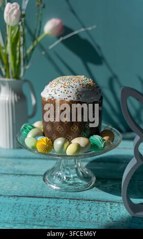 Traditionelles Paska- oder Pasca-Osterbrot, das auf einem gläsernen Kuchenständer mit dekorativen Ostereiern und einem Hasenornament vor einem blauen Holz-Hintergrund präsentiert wird Stockfoto