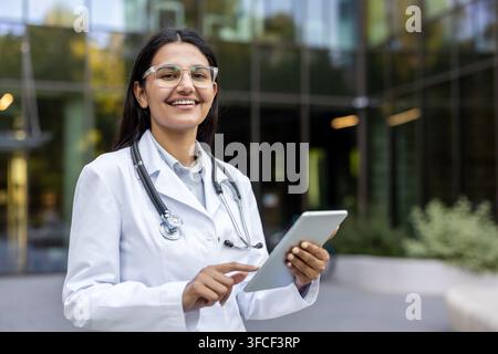 Indische Ärztin in weißem Kittel und Stethoskop, die im Freien lächelt, ein digitales Tablet hält und gestikuliert, repräsentiert moderne Telemedizin, medizinische Technologie und berufliche Expertise Stockfoto