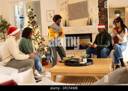 Verschiedene Familie trägt Weihnachtsmützen und Pullover, sitzt am Kamin und tauscht Geschenke in der Nähe des Baumes aus Stockfoto