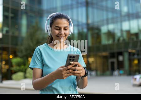 Junge Frau lächelt, während sie Kopfhörer trägt und ein Smartphone benutzt, um sich mit digitalen Inhalten und Kommunikation im Freien in einer städtischen Umgebung zu verbinden Stockfoto