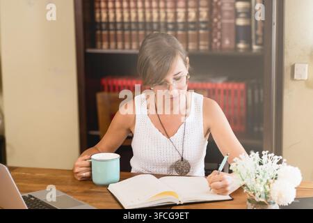 Junge Frau mit Brille schreibt in ein offenes Notizbuch an einem Holztisch mit Laptop, Kaffeetasse und Blumen in einem gemütlichen Arbeitszimmer zu Hause. Stockfoto