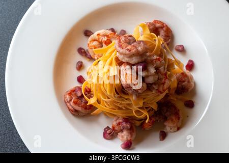 Oberansicht eines goldenen Fettuccine-Nests mit rosa Garnelen und rubinroten Granatapfelsamen, die auf einem breiten weißen Teller verstreut sind, dunkle graue Oberflächenecke Stockfoto