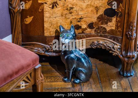 Eine glänzende schwarze Keramikkatzenfigur mit grünen Augen sitzt vor einem Möbelstück mit floraler Holzmarqueterie im Musée de l'École de Nancy in der Rue du Sergent Blandan in Nancy, Grand Est, Frankreich. Stockfoto