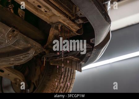 Schmutzige Fahrzeugunterseite mit rotem Flüssigkeitstropfen aus dem unteren Motorbereich. Rad, Spritzschutz und Metallstützen sind im Workshop im Nahaufnahme sichtbar Stockfoto