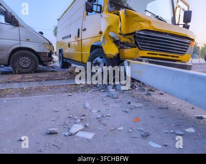 Niedrigwinklige Außenaufnahme zeigt die Nachwirkungen eines Verkehrsunfalls. Zwei beschädigte Vans, einer hellfarben und einer gelb, sind von Trümmern auf der Straße umgeben Stockfoto