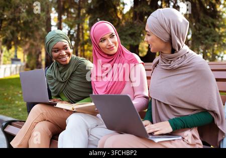 Drei Moderne Isamico-Damen Mit Laptops Und Büchern Lernen Sie Gemeinsam Sprechen, Sitzen Sie Auf Der Bank Im Park Im Freien. E-Learning, Studentenleben Stockfoto