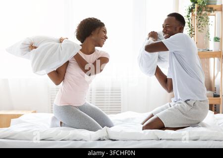 Fröhliche liebende afroamerikanische Paar kämpfen mit Kissen im Bett, Spaß zusammen zu Hause, leeren Raum. Glücklich schwarz Mann und Frau im Schlafanzug hav Stockfoto