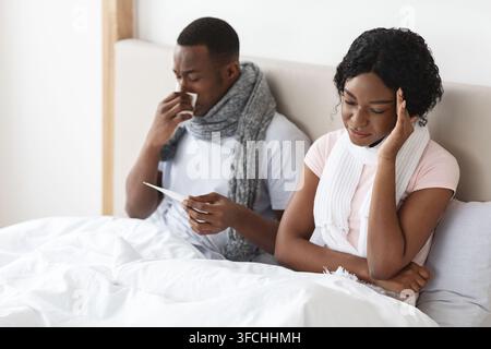 Krankes afroamerikanisches Paar, in Tücher gewickelt, sitzt im Bett, misst Fieber und niesende Nasen. Junge schwarze Familie von zwei ist krank geworden, bleib Stockfoto