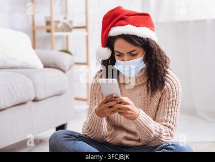 Allein feiern. Unzufriedene schwarze Dame, die ein Smartphone benutzt und auf dem Boden im Wohnzimmer sitzt. Verwirrte afroamerikanische Frau, die eine medizinische Maske trägt und s Stockfoto