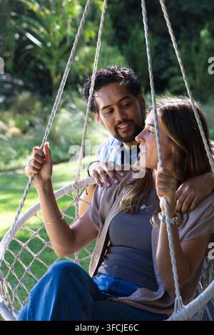 Verschiedenes Paar sitzt auf einem Makramee-Hängeswing im Hinterhof, beide tragen Jeans und halten Seile Stockfoto