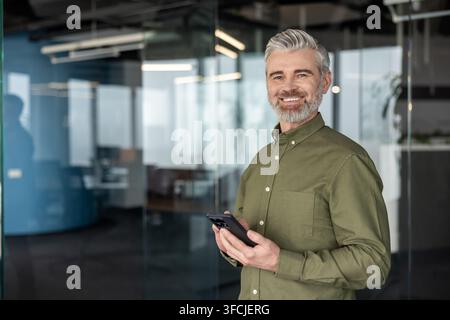 Selbstbewusster reifer Geschäftsmann mit grauen Haaren und Bart, der in einem modernen Büro steht, in die Kamera lächelt und ein Smartphone hält, was Professionalität, Führungskompetenz und digitale Vernetzung vermittelt Stockfoto