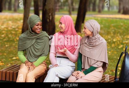 Drei Muslimische Junge Frauen Im Gespräch Sitzen Auf Der Bank Und Verbringen Ein Wochenende Im Park Mit Hijab-Kopftuch. Glückliche Islamische Studentinnen Genießen Es Stockfoto