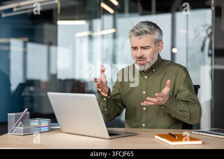 Reifer Mann fühlt sich gestresst und verwirrt, gestikuliert aktiv mit den Händen, während er auf einen Laptop-Bildschirm schaut, und steht vor technologischen Problemen in einem modernen Büroumfeld Stockfoto
