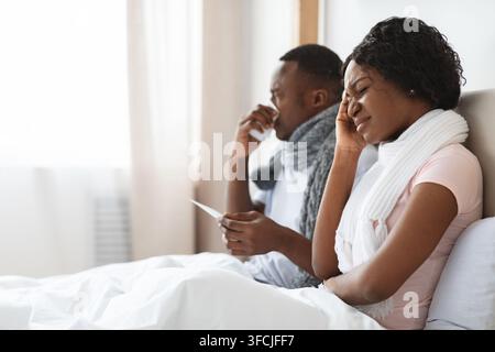 Kranke afroamerikanische Männer und Frauen, in Tücher gewickelt, sitzen im Bett, messen Fieber und niesende Nasen. Die junge schwarze Familie von zwei Leuten hat einen Cor bekommen Stockfoto